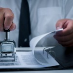 Business approval and certificate concept. Close-up of person's hand stamp with certification stamp on document. At desk, lawyer or businessman document signing.